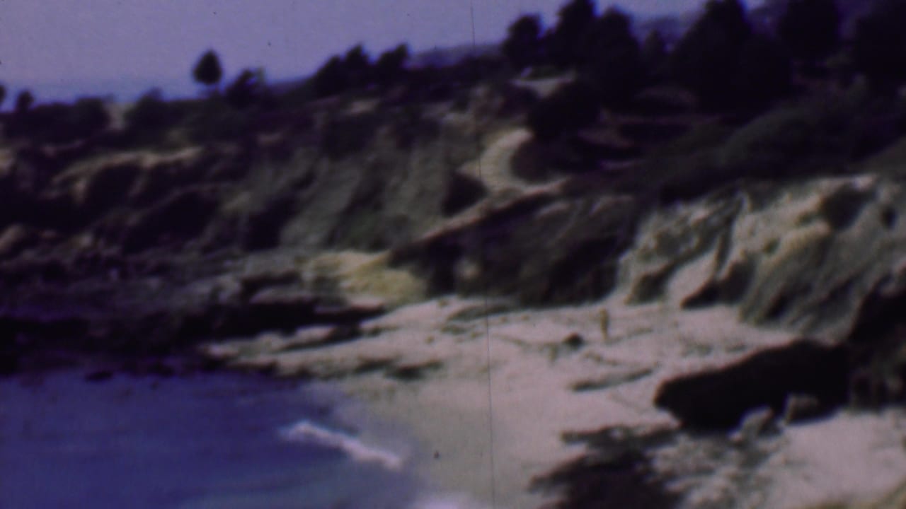 1961 San Diego coastal home-movie frame showing a rocky sandy cove, blue water, eroded bluffs, and dark trees along the ridge.