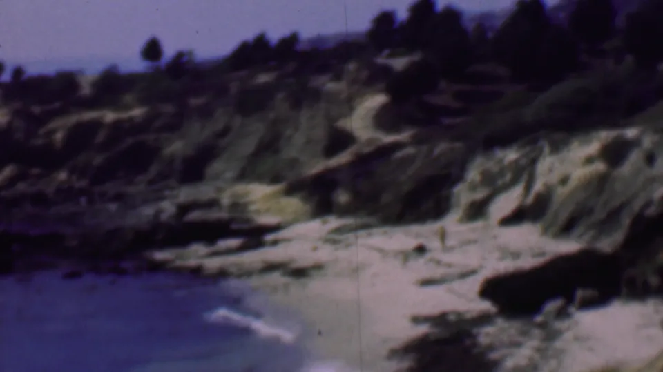 1961 San Diego coastal home-movie frame showing a rocky sandy cove, blue water, eroded bluffs, and dark trees along the ridge.