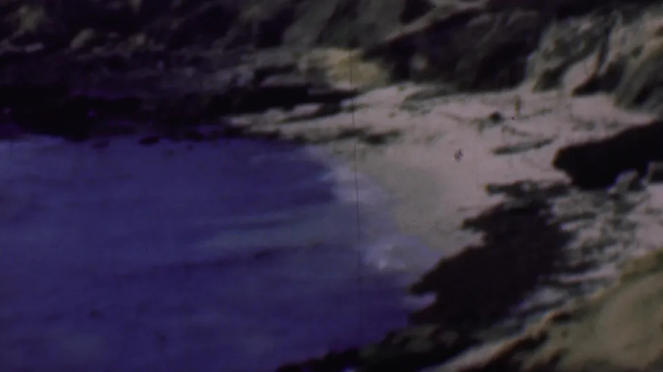 1961 San Diego shoreline view with a curved sandy beach, calm blue water, dark rock outcrops, and pale bluff faces above the cove.