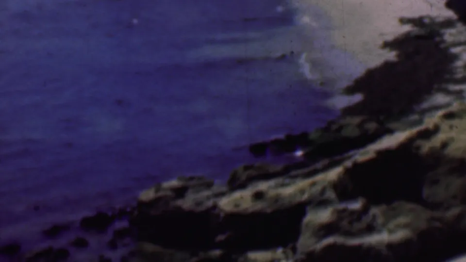 1961 San Diego home-movie frame looking across blue cove water toward rocks, pale sand, and a dark shoreline edge beneath coastal bluffs.