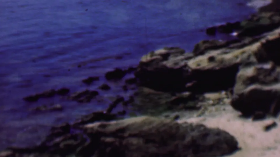 1961 San Diego coastal frame showing blue water, scattered dark rocks, a pale sandy edge, and rugged shoreline formations in soft 8mm color.