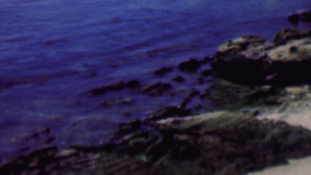 Soft 1961 8mm frame of rocks beside the ocean at a San Diego beach, with deep blue water and a sandy edge partly visible.