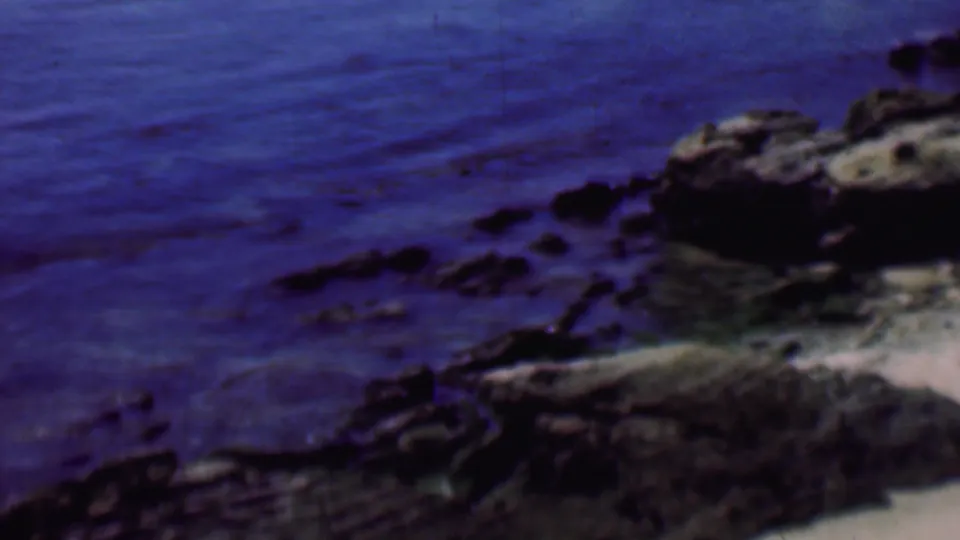 Soft 1961 8mm frame of rocks beside the ocean at a San Diego beach, with deep blue water and a sandy edge partly visible.