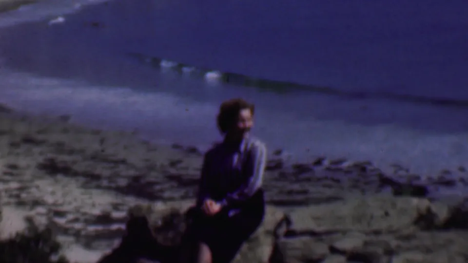 A woman in a striped blouse and dark skirt sits on a rocky outcrop beside the beach in supported 1961 San Diego home-movie footage.