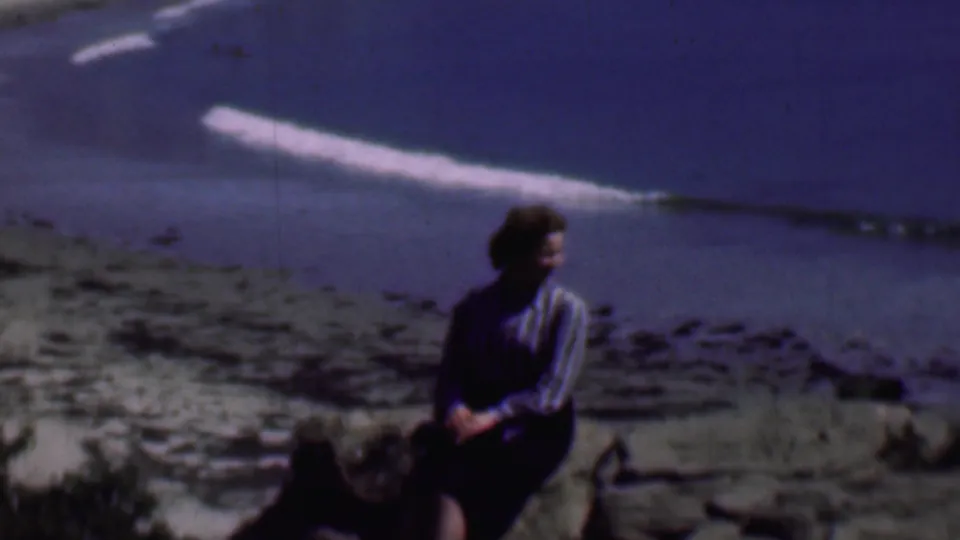 The woman sits in profile on coastal rocks while small waves roll onto the San Diego beach in soft, color-shifted 8mm footage from 1961.