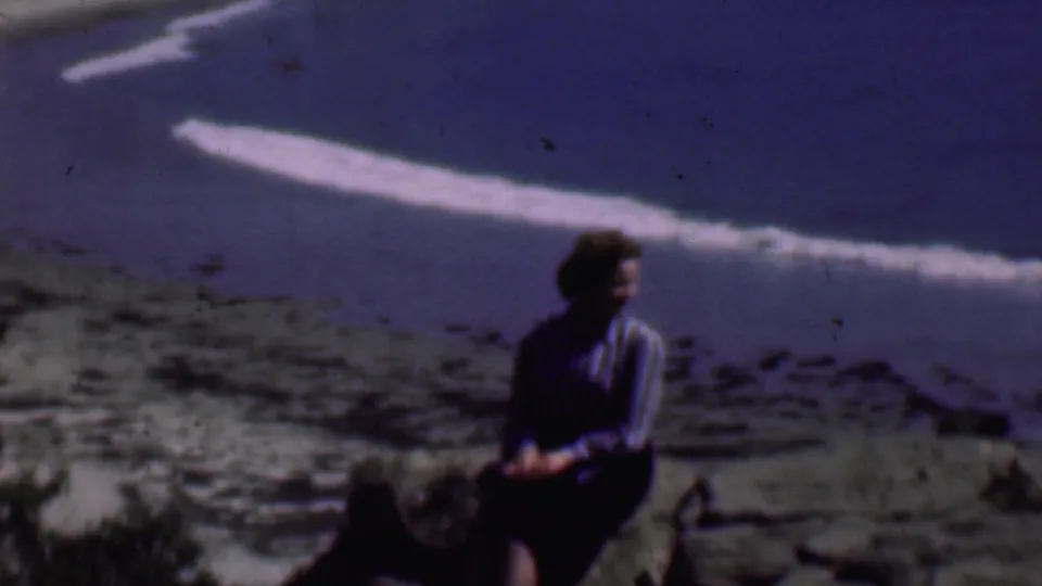 A seated woman remains on the rocky shoreline as a bright band of surf crosses the beach behind her in 1961 San Diego home-movie footage.