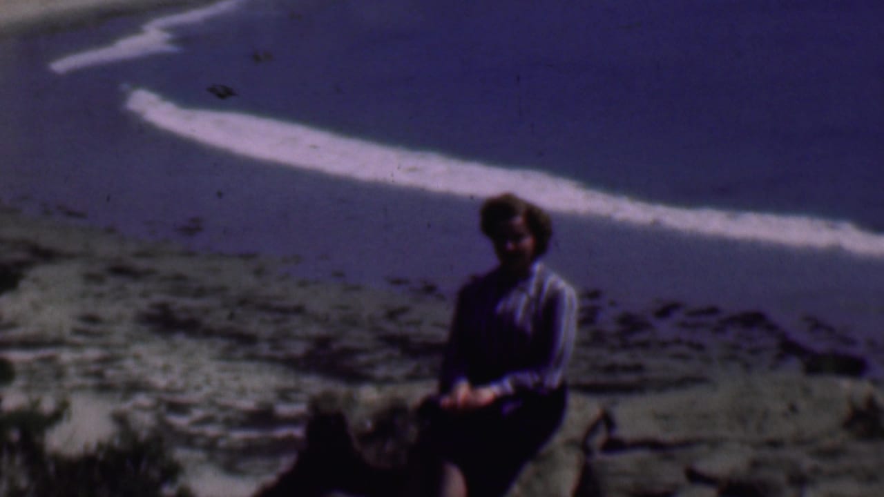 In supplied 1961 San Diego home-movie footage, a woman in a striped blouse and dark skirt sits on a large rock overlooking a rocky beach and ocean surf.