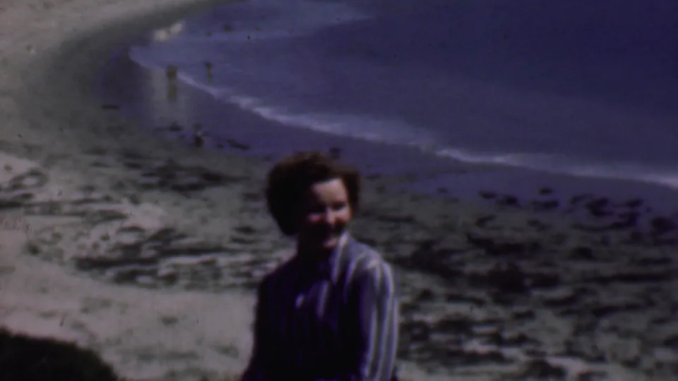 The woman turns toward the camera and smiles, with the rocky San Diego beach and pale surf line visible behind her in soft 8mm color.