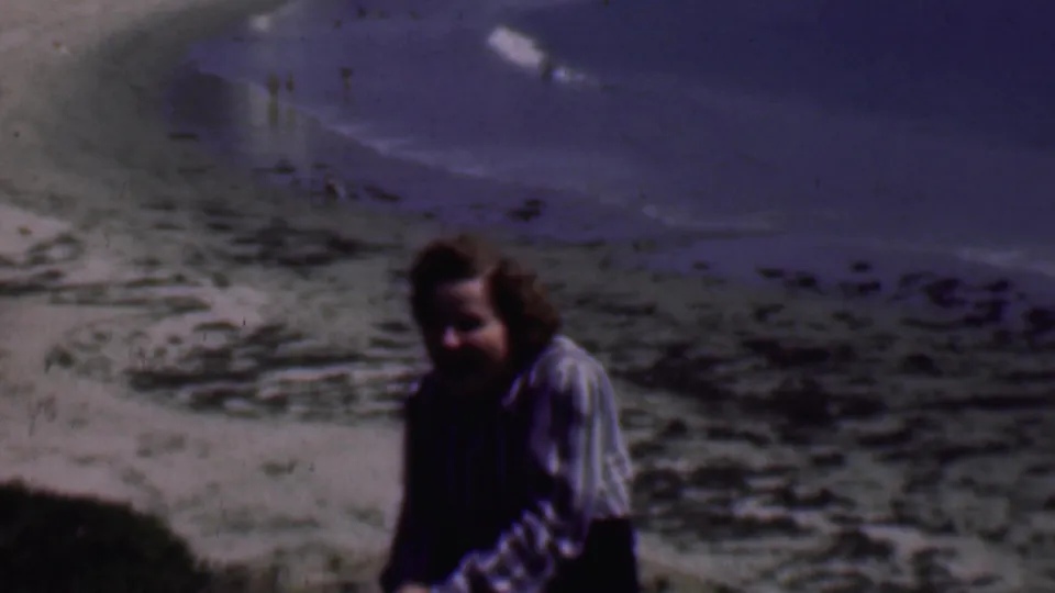 The woman remains seated in partial shadow as the handheld camera frames her against the rocky shoreline and small waves below.