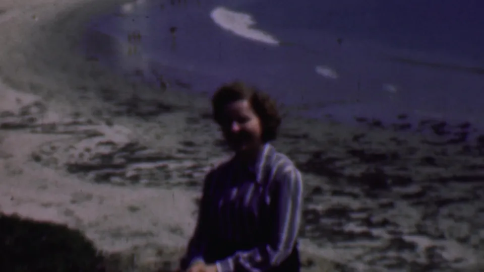 A woman in a striped blouse smiles from a coastal rock overlook, with faded 8mm color showing ocean water, scattered rocks, and beach texture behind her.