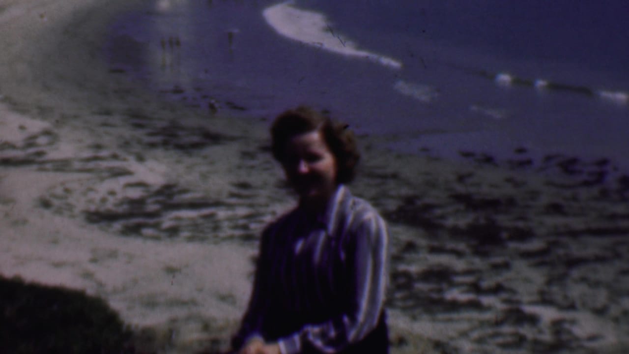 In 1961 San Diego home-movie footage, an adult woman sits on a sandy beach near the waterline with blue ocean and pale surf in the background.