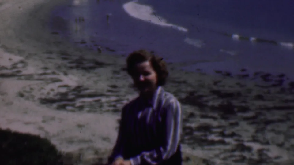 In 1961 San Diego home-movie footage, an adult woman sits on a sandy beach near the waterline with blue ocean and pale surf in the background.