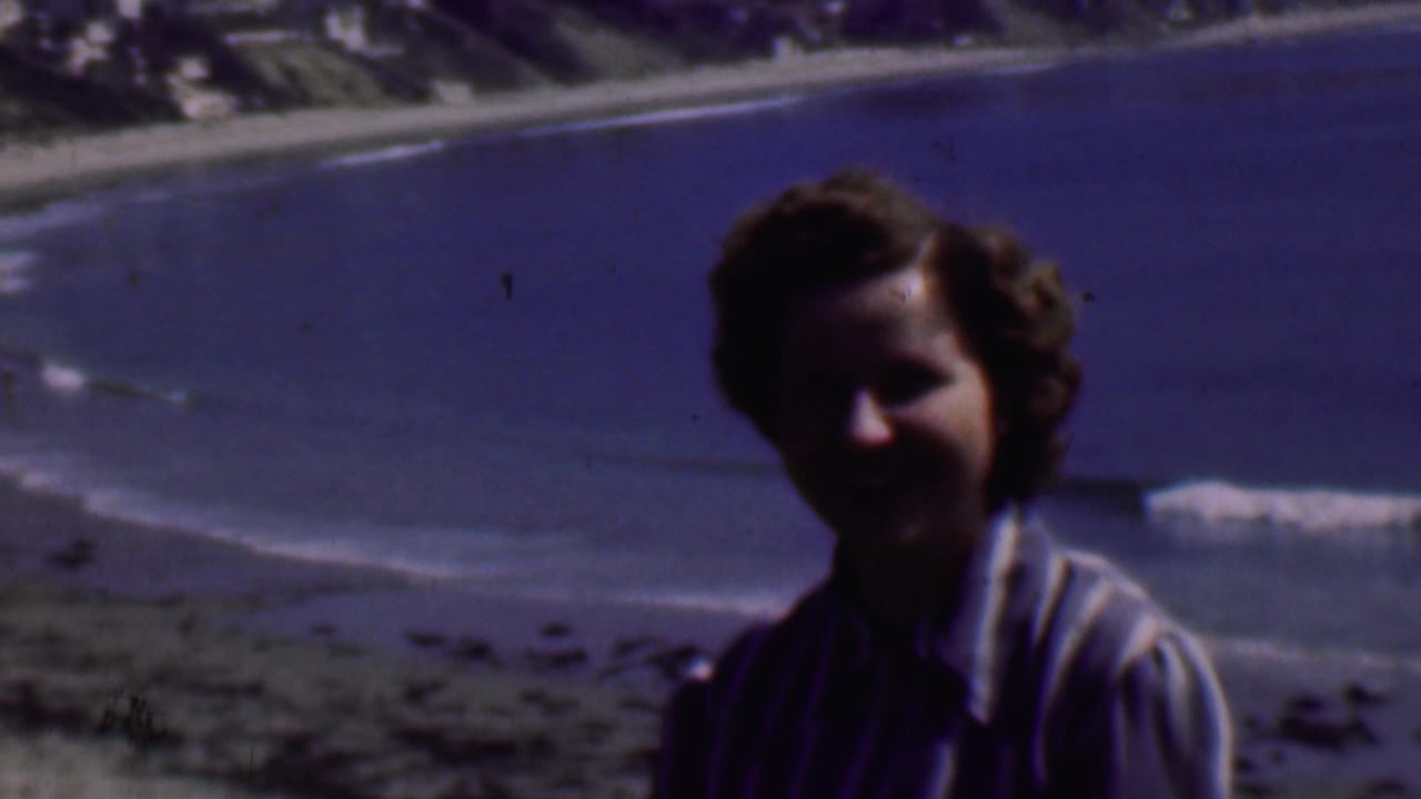 A smiling adult woman in a striped collared shirt faces the camera on a San Diego beach, with blue water and small waves behind her.