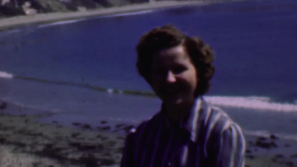 The camera holds on the seated woman as she looks toward the lens, with the beach, rolling surf, and distant shoreline visible behind her.