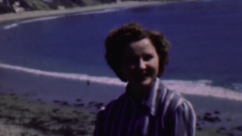 A smiling adult woman remains seated in the foreground of the 1961 San Diego beach scene, with pale surf and blue ocean filling the background.