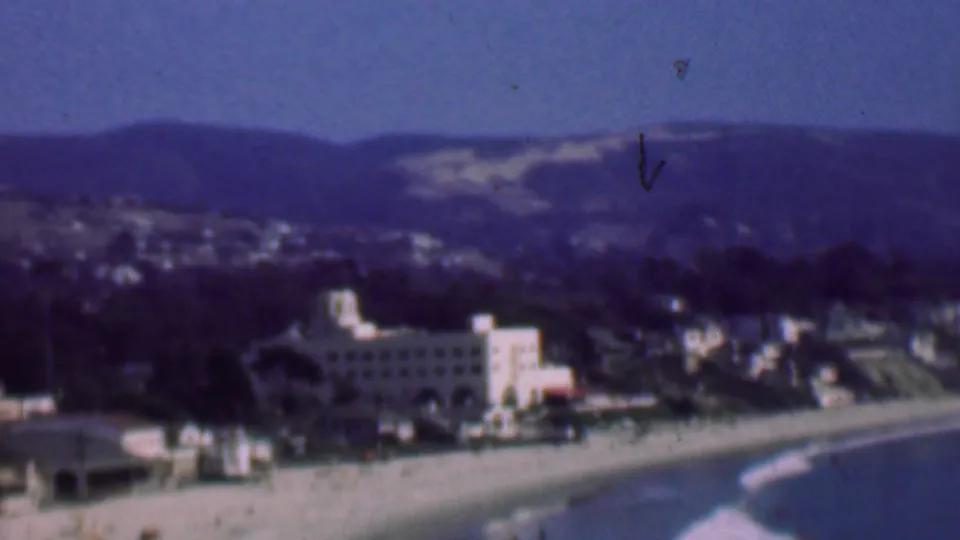 1961 San Diego home-movie frame showing a distant coastal overlook with a sandy beach, large beachfront building, surf line, town structures, and rolling hills.