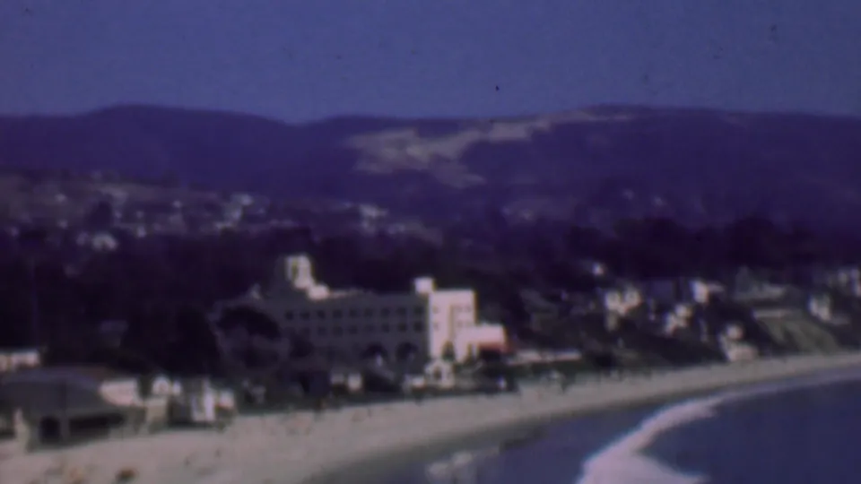 1961 San Diego home-movie frame of the coastline from an overlook, with a curved beach, prominent beachfront building, distant town, and hills beyond.