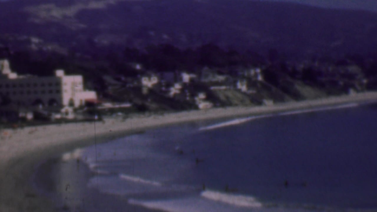 1961 San Diego coastal home-movie frame showing a curving sandy beach, small waves, hillside residences, and a prominent white building near the shore.