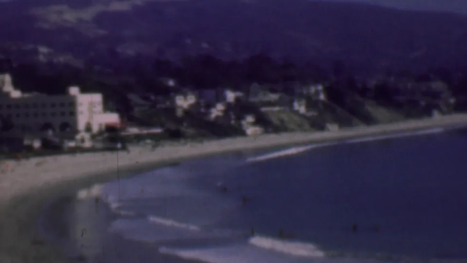 1961 San Diego coastal home-movie frame showing a curving sandy beach, small waves, hillside residences, and a prominent white building near the shore.