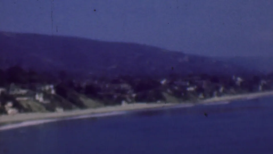 Soft 8mm frame of the 1961 San Diego shoreline, showing hillside residences above a beach with thin white surf lines and hazy hills beyond.