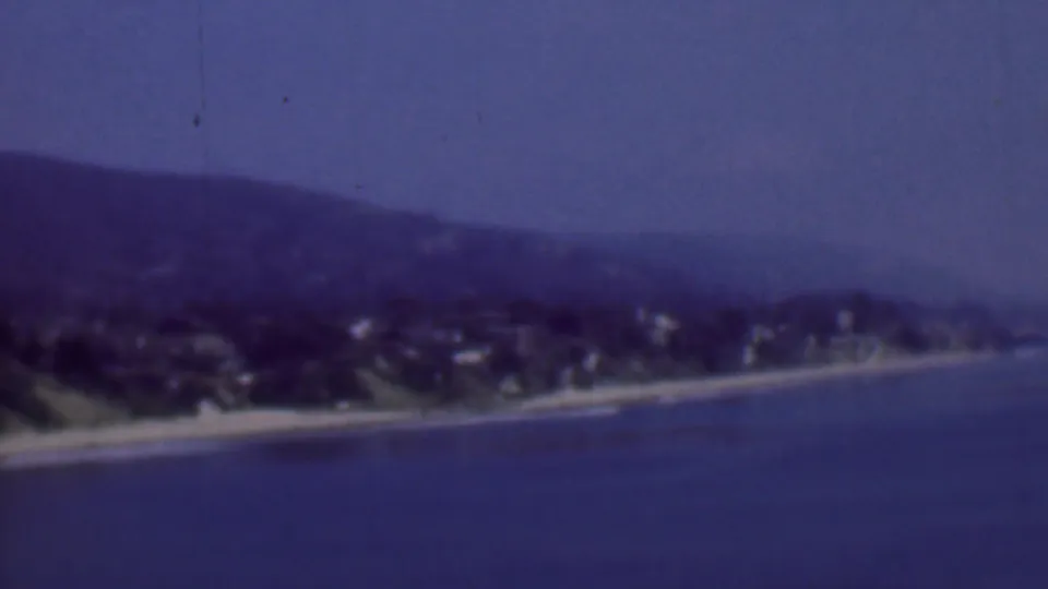 Wide 1961 San Diego beach panorama with blue water in the foreground, a narrow shoreline, hillside homes, and hazy terrain in the background.