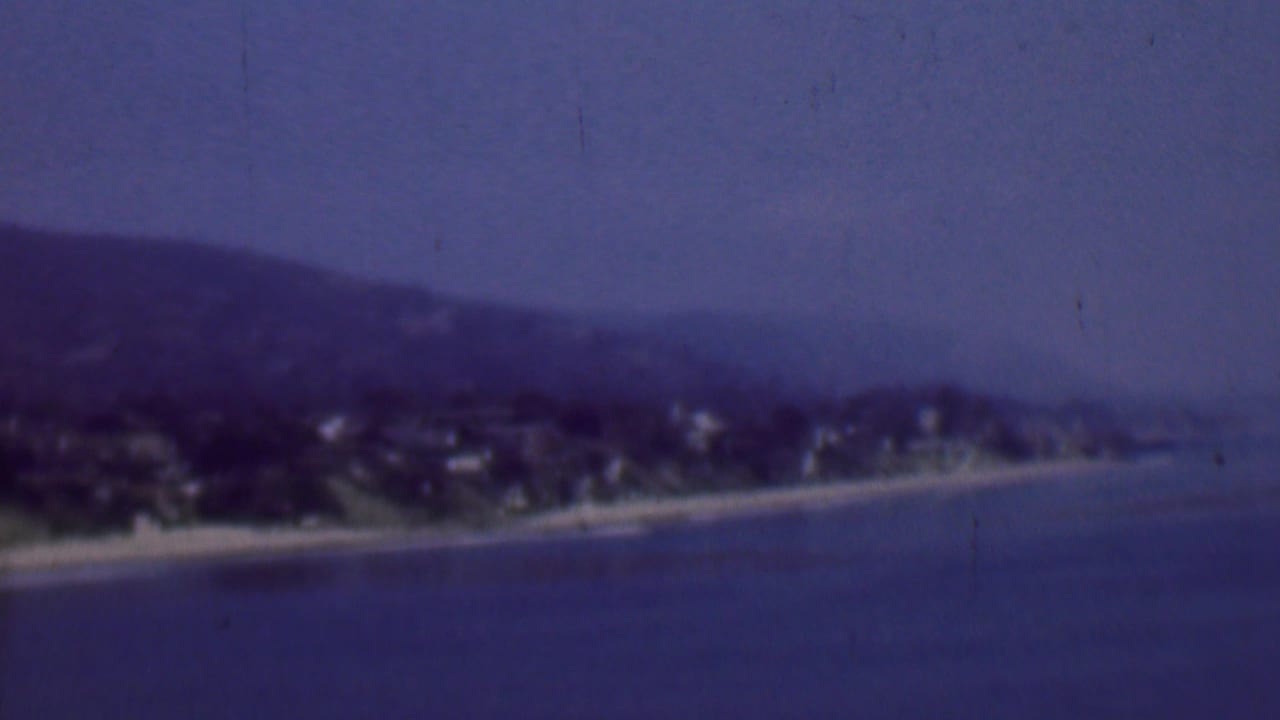 1961 8mm view of a San Diego-area coastline with a sandy beach, calm sea, hillside housing, and soft haze across the frame.
