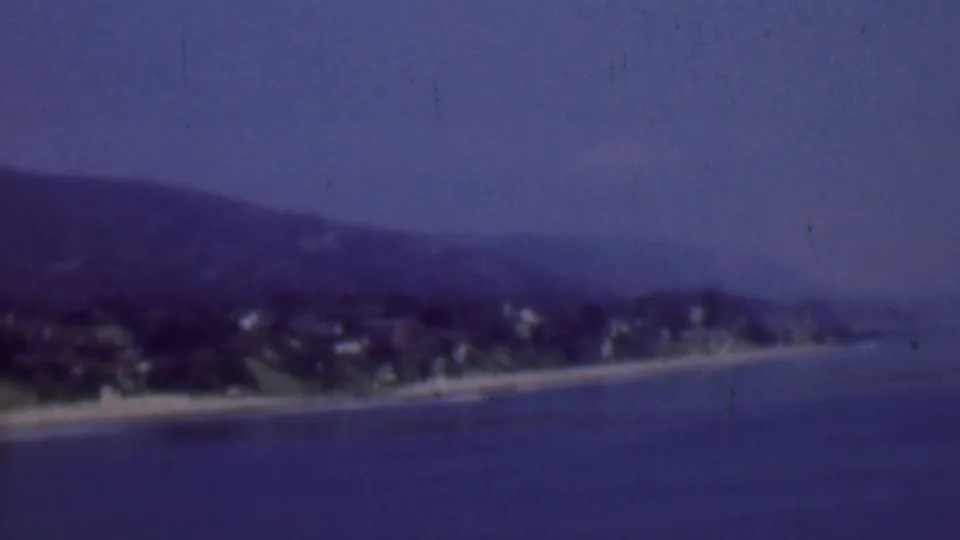 1961 8mm view of a San Diego-area coastline with a sandy beach, calm sea, hillside housing, and soft haze across the frame.