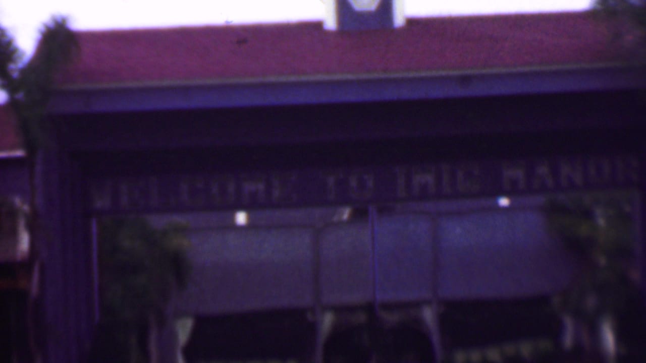 Soft 8mm frame of a building entrance in the San Diego-area footage, showing a red roofline, palms, shaded awnings, and a partially legible welcome sign.