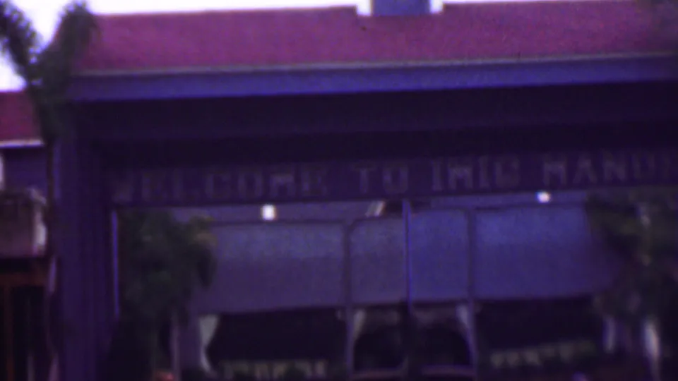 1961 8mm home-movie view of a red-roofed building exterior with palm trees, blue-toned awnings, and a faded sign that appears to begin with welcome.