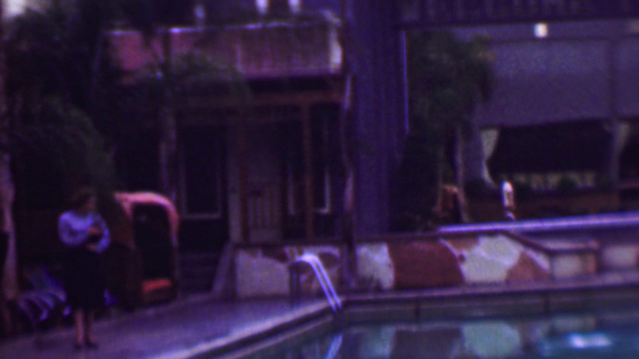 In 1961 San Diego home-movie footage, a woman in a light blouse and dark skirt stands near a swimming pool beside lounge chairs and palm trees.