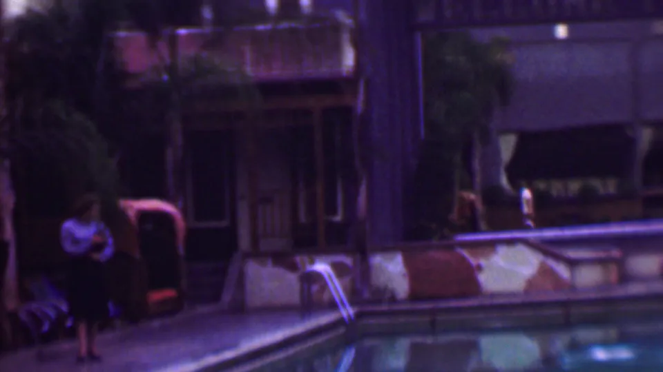 In 1961 San Diego home-movie footage, a woman in a light blouse and dark skirt stands near a swimming pool beside lounge chairs and palm trees.