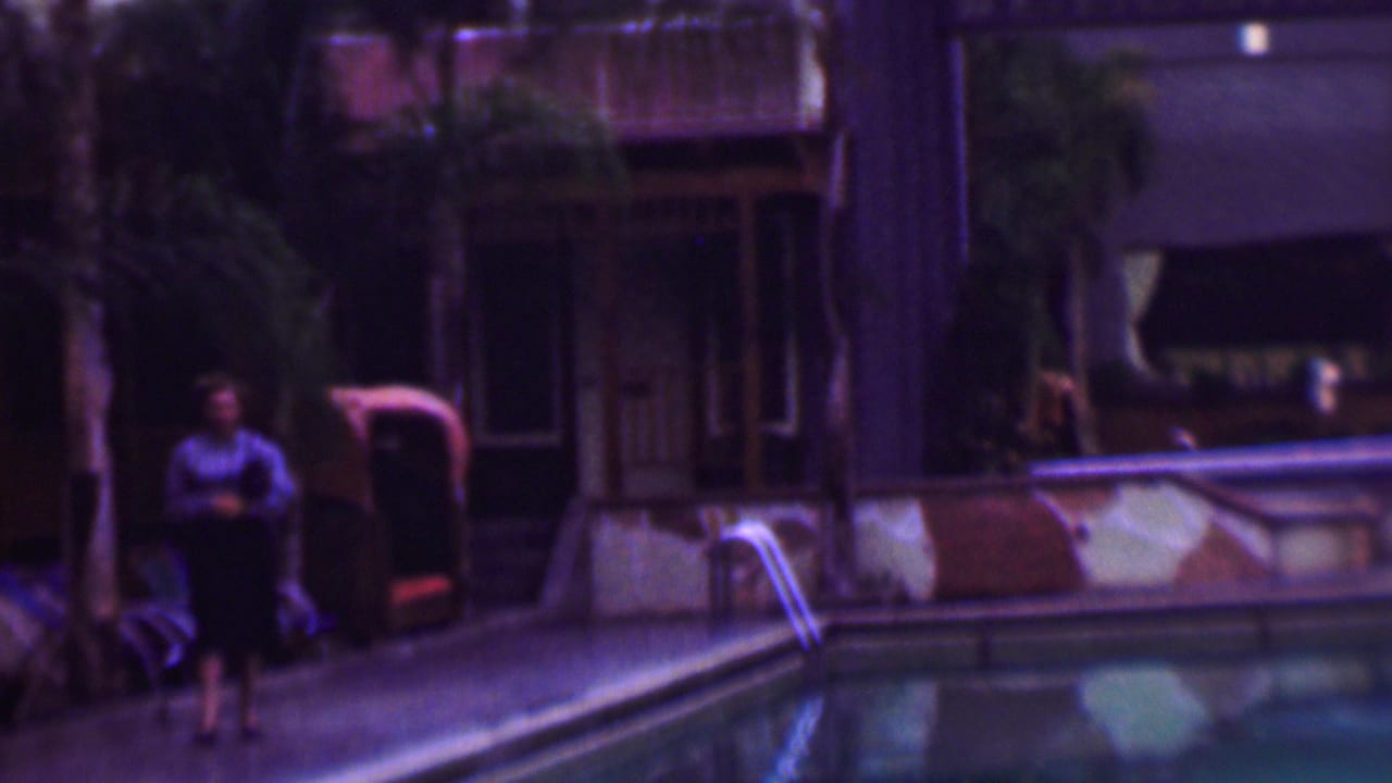 A woman walks along a San Diego poolside patio in 1961, with a metal pool ladder, lounge seating, palms, and a wooden building in the background.