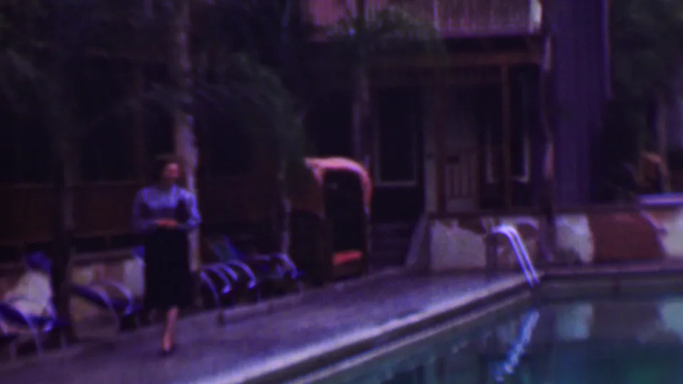 Near the end of the clip, the woman walks along the poolside patio as lounge chairs, palms, a metal ladder, and a wooden building remain in view.