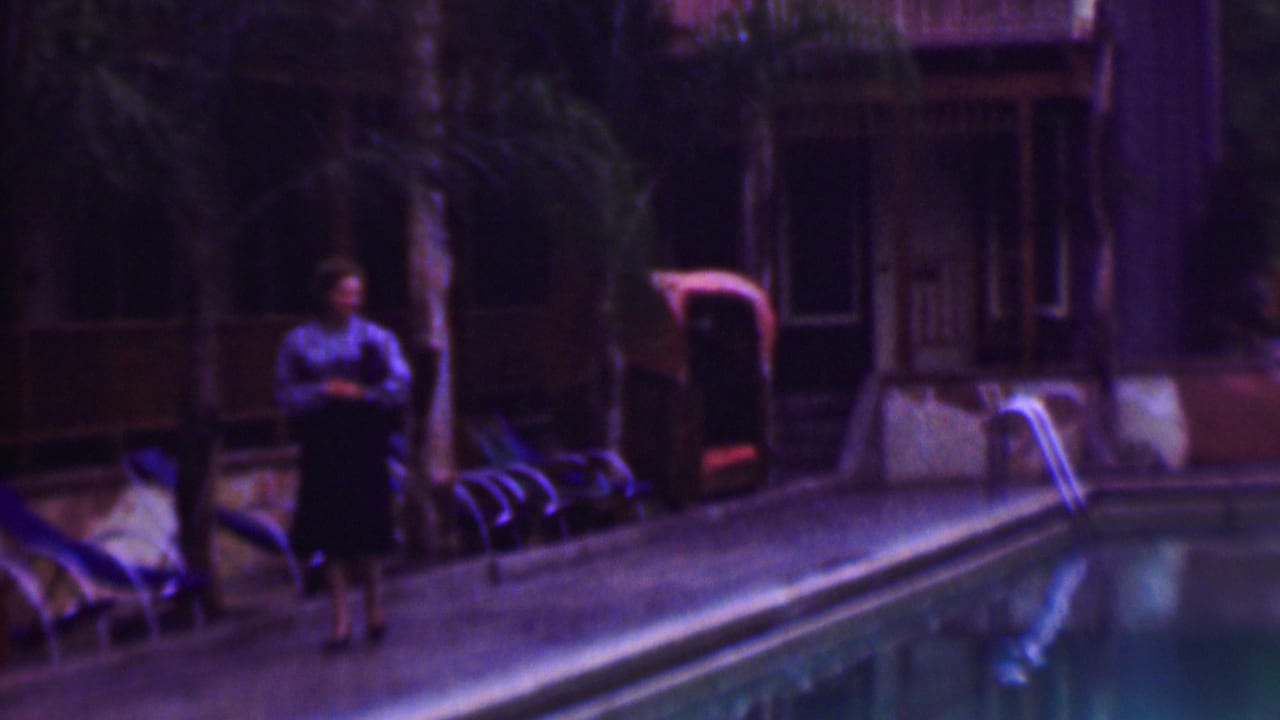 A woman in a blouse and dark skirt stands by a large pool in supplied 1961 San Diego home-movie footage, with lounge chairs, palm trees, and a building in the background.