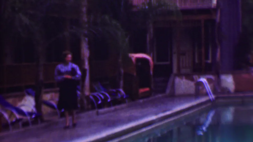 A woman in a blouse and dark skirt stands by a large pool in supplied 1961 San Diego home-movie footage, with lounge chairs, palm trees, and a building in the background.