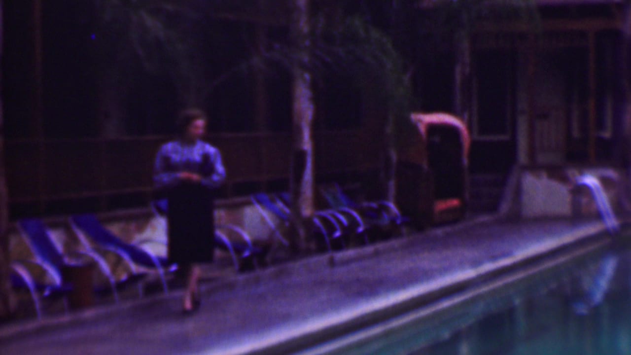 Woman walking along the pool deck past lounge chairs and palm trees.