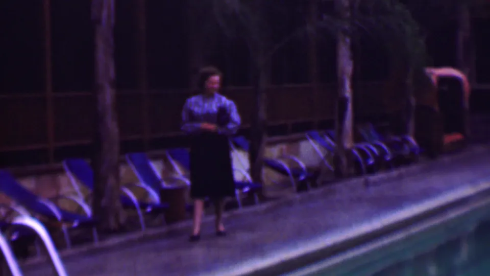The woman appears nearer on the walkway beside the large pool, with stacked lounge chairs, palms, and the dim hotel exterior behind her.