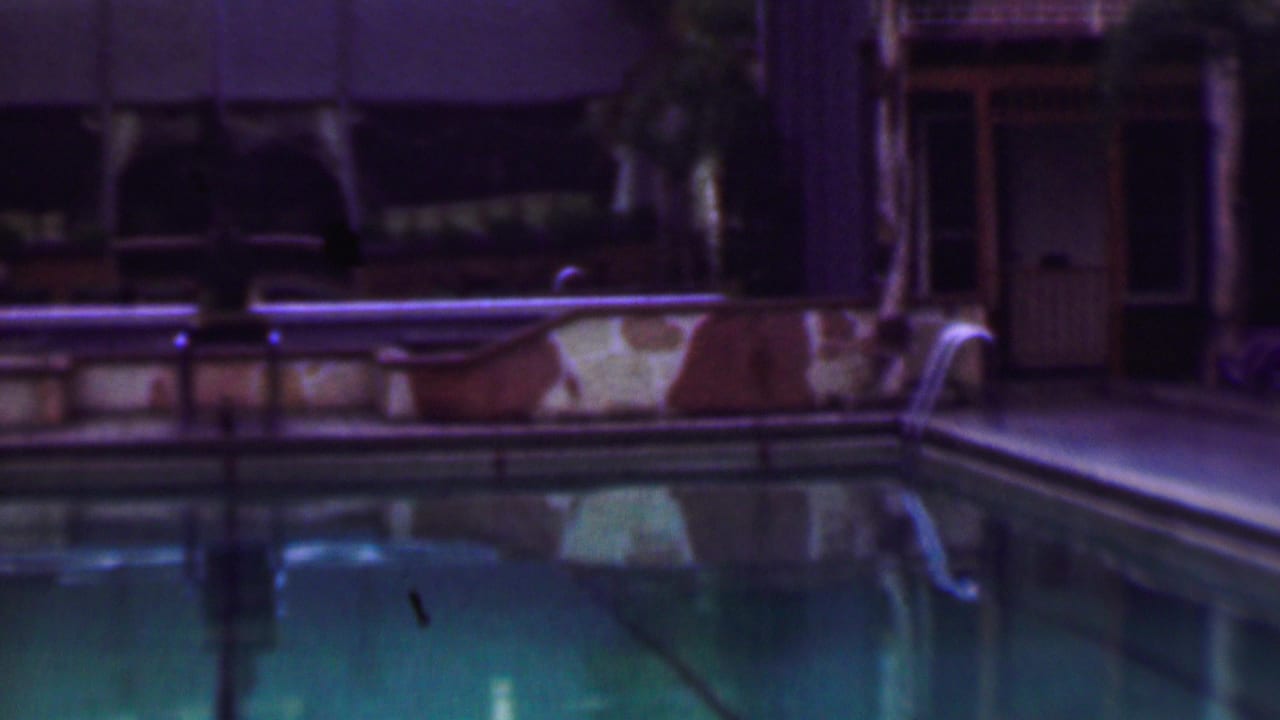 Night pool view with reflective water, railings, stone border, and a building entrance at the right.