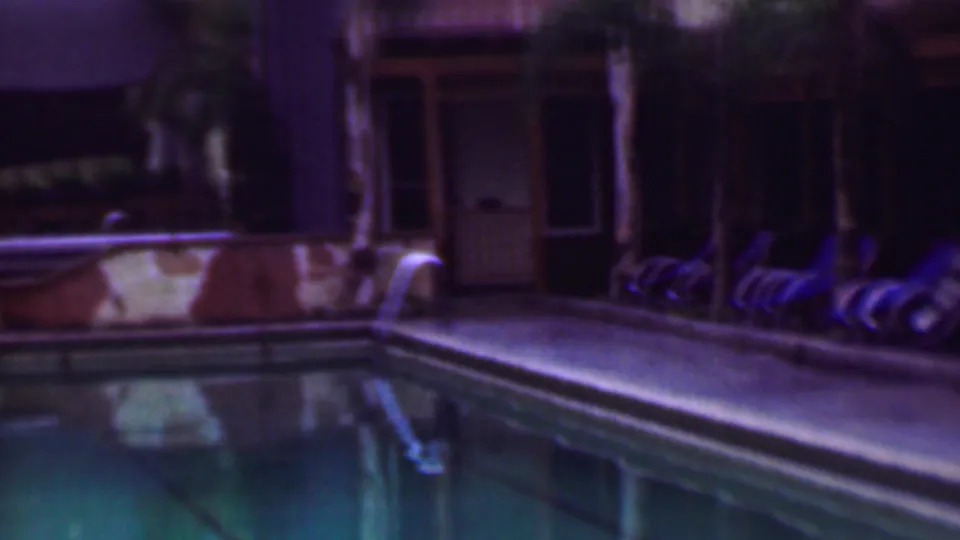 1961 San Diego frame showing a pool corner at night, with turquoise water, a pale deck, a handrail, and a row of lounge chairs near a shadowed building.
