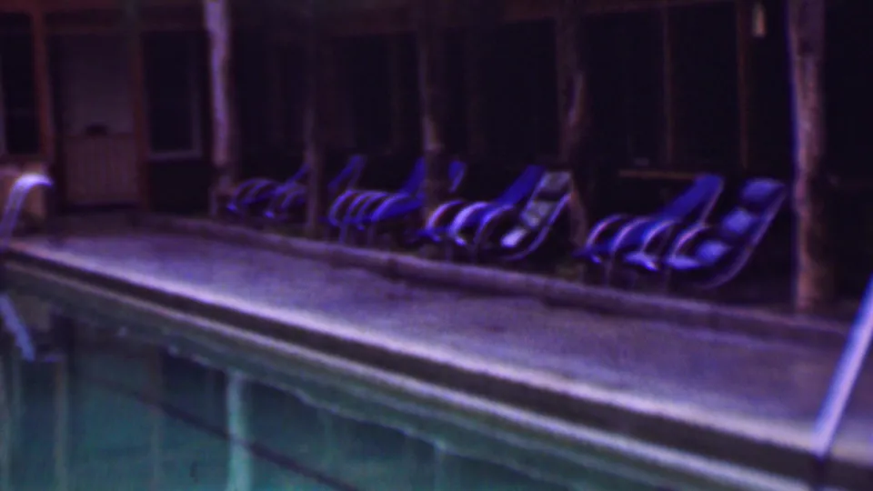 1961 San Diego home-movie frame with blue lounge chairs lined along a nighttime pool deck, framed by wooden posts, dark windows, and reflective pool water.