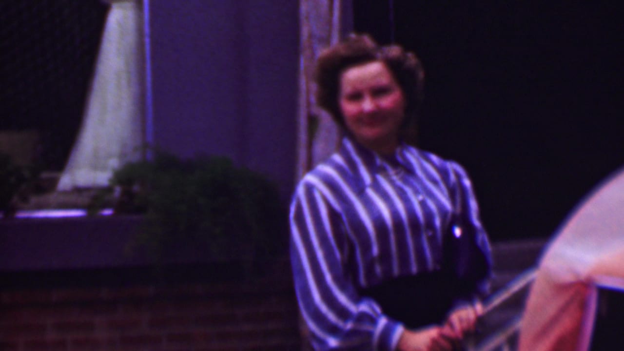 1961 San Diego home-movie frame with an adult woman in a blue-and-white striped blouse smiling near the poolside, with an umbrella or parasol and patio plants behind her.