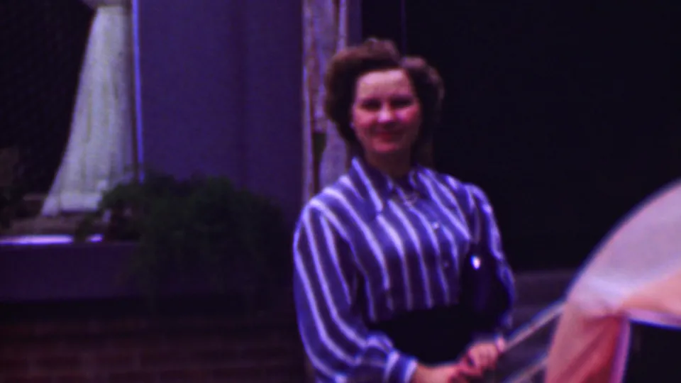 1961 San Diego home-movie frame showing an adult woman in a striped blouse smiling toward the camera near a pool, with a pale umbrella or parasol and patio background visible.