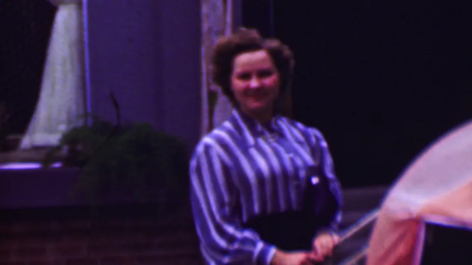 1961 San Diego home-movie frame of an adult woman posing by a swimming pool, with brickwork, potted greenery, and a pale umbrella or parasol in the dim patio setting.