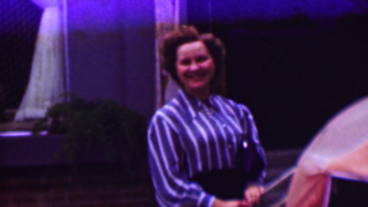 A woman in a blue-and-white striped blouse and dark slacks smiles toward the camera in 1961 home-movie footage from the United States, with porch details and a railing behind her.