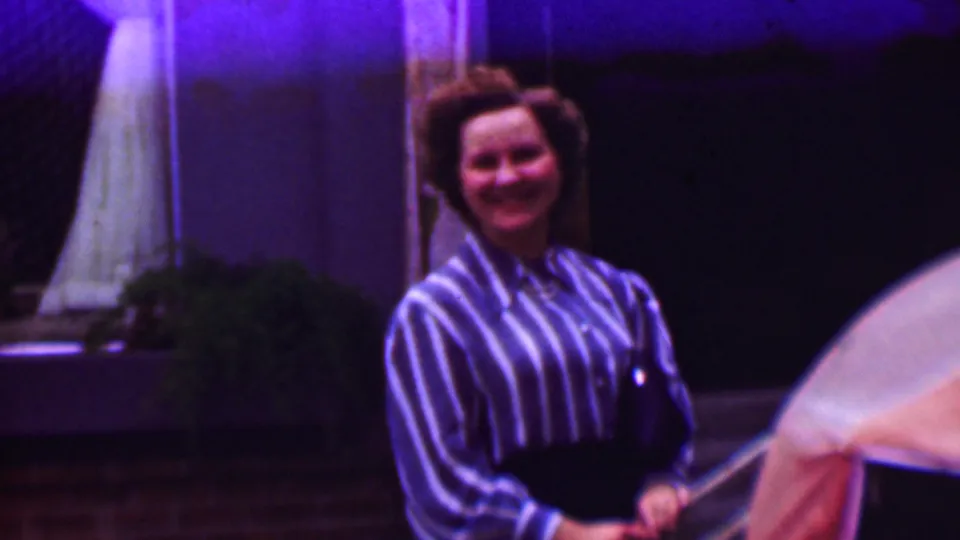 A woman in a blue-and-white striped blouse and dark slacks smiles toward the camera in 1961 home-movie footage from the United States, with porch details and a railing behind her.