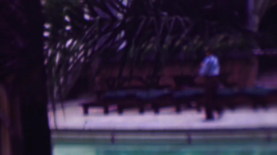 A man in a light blue shirt and dark pants walks through a poolside area with lounge chairs, water, and tropical planting visible in soft 8mm focus.