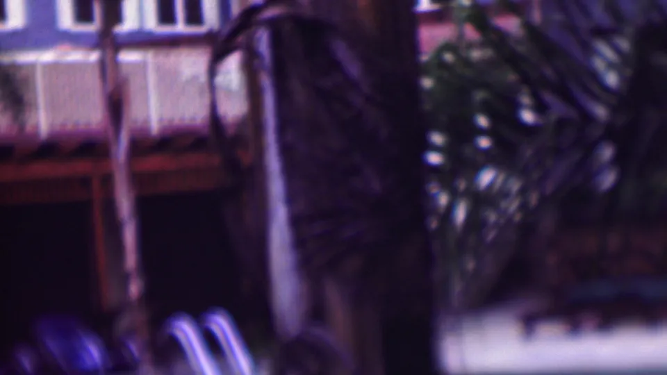1961 home-movie frame of a shadowed pool area with palm trunks, white balcony rails, a red-toned overhang, and blue lounge chairs below.