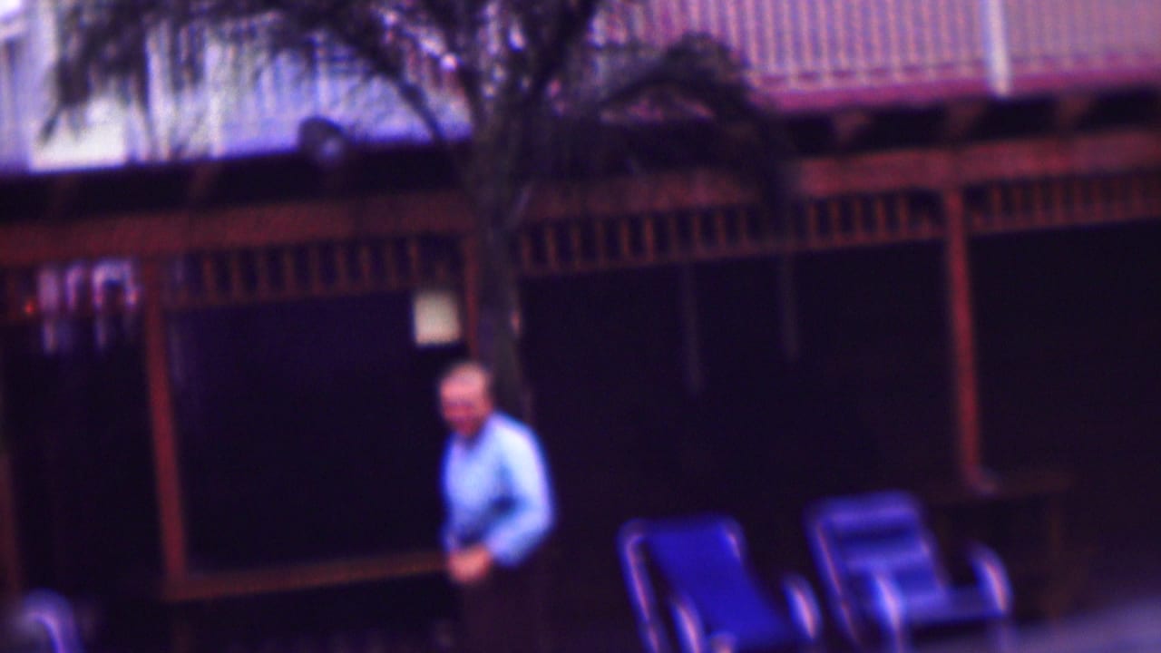 In supported 1961 San Diego home-movie footage, an adult man in a light blue shirt stands near blue deck chairs in front of a shaded open-air porch.