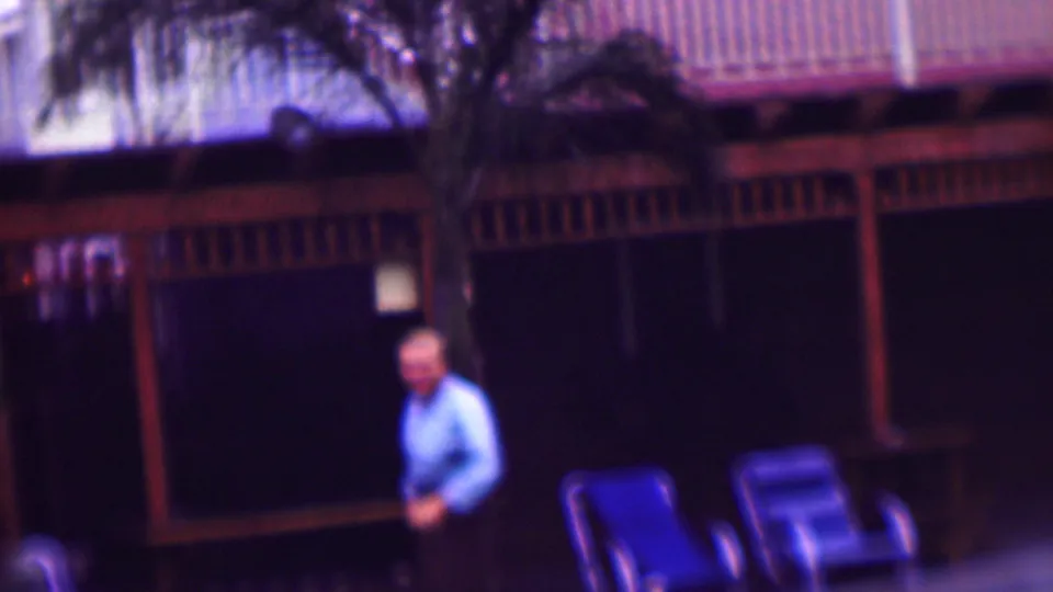 In supported 1961 San Diego home-movie footage, an adult man in a light blue shirt stands near blue deck chairs in front of a shaded open-air porch.