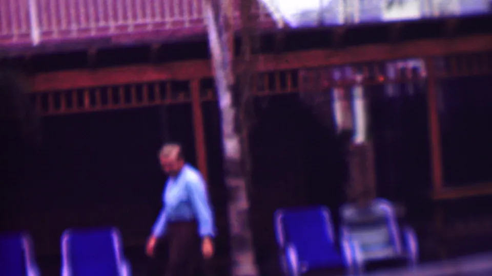 The man continues walking through the outdoor area, passing blue deck chairs and a dark open-air porch with wood railings.